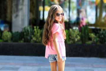 Fashion kid summer in the city stands on a streetl. Trendy girl in sunglasses standing on the street at sunset.
