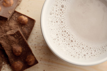 White cup with hot cocoa stands on wooden table near pieces of chocolate with nuts sprinkled with cocoa powder