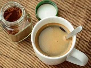 Photo of coffee in a white cup and spoon standing near the jar of coffee and green cover. Making aromatic coffee