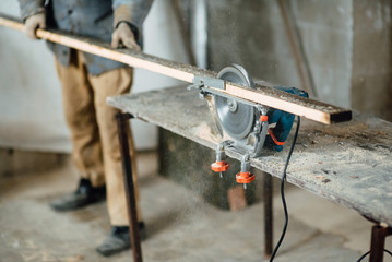 man in work clothes by profession carpenter handles wooden board on circular saw on a wooden workshop table, joiner works on machine in factory