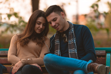 Young couple listening to music,smiling,looking at the phone and sitting on a bench in park.