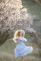 A young long-haired blonde is walking in a blooming garden. Dress and hair fluttering like in a fairy tale