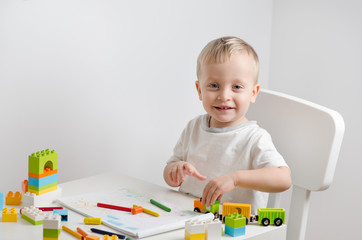 Little blond child playing with lots of colorful plastic blocks, with a toy train and draws at the table. Little boy sits on a chair at the white table.