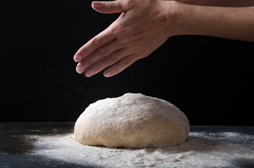 Female hands sprinkling flour over fresh dough on dark background.