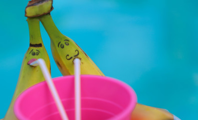 Two bananas with painted faces are drinking from a pink glass of juice through a straw on an inflatable mattress in the pool. Humor