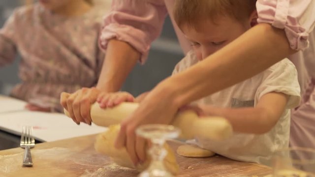 The mother together with her little baby son rolls the kneaded dough using rolling pin. She stands over him and he sits on the chair trying to put his little hands on the rolling pin.