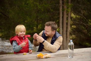 Little boy and his father eating snacks during picnic on coast of mountain river in Switzerland.