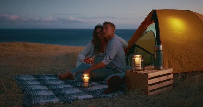 Attractive couple snuggling with candles and tent in beautiful fields at dusk, camping romance, adventure