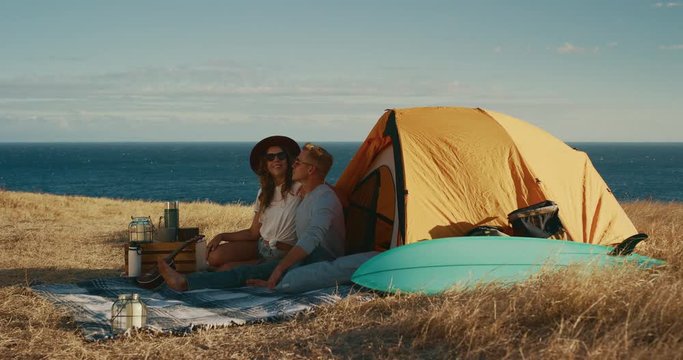 Attractive couple kissing in comfortable campsite surrounded by golden fields and ocean, summer surf camping adventure