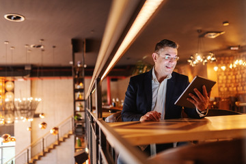 Smiling Caucasian middle aged man dressed smart casual using tablet and sitting in cafeteria while waiting for coffee.
