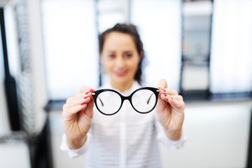 Woman at optician holding eyeglasses she want to buy. Selective focus on glasses. - obrazy, fototapety, plakaty