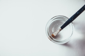 Blue Paintbrush With Gold Bristles Resting In A Glass Jar of Water Next To A Blank Canvas