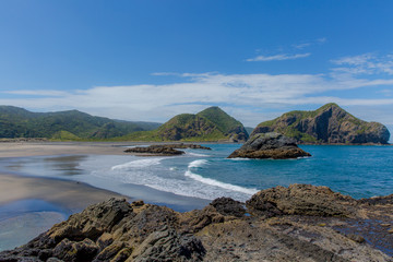 View of the Whatipu beach from the Ninepin Rock to the Paratutae Island. White waves on the beach and rock formations, summer day.