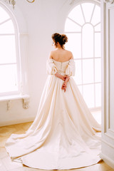 Elegant young woman in creamy satin dress standing near the window
