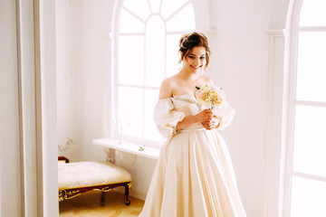 Elegant young woman in creamy satin dress standing near the window