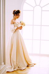 Elegant young woman in creamy satin dress standing near the window in the chapel before getting married