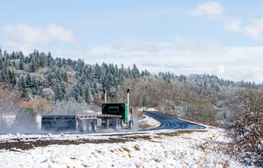 Green classic big rig semi truck with empty flat bed semi trailer moving on the wet winter snow road with snowy trees on backgroud
