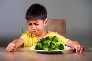 7 or 8 years old upset and disgusted Asian kid sitting on table in front of broccoli plate looking...