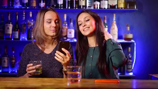 two pretty young women are dancing near bar counter and viewing pictures in smartphone