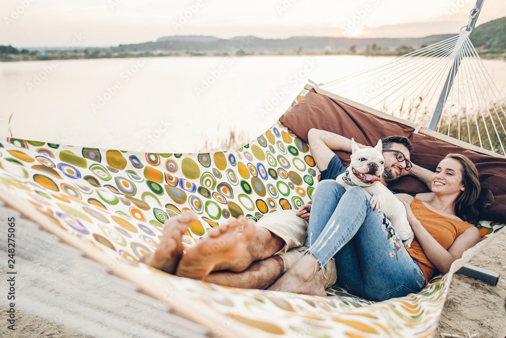 Wall mural hipster family on vacation concept, happy woman and man relaxing on a hammock at the beach with thei