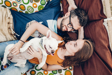 Happy young family portrait, cute stylish woman holding happy bulldog near handsome man in sunglasses while relaxing on a hammock on the beach
