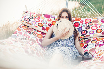 Beautiful hipster girl in stylish dress hiding behind hat and smiling while resting on a hammock on...