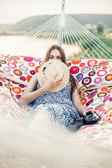 Beautiful hipster girl in stylish dress hiding behind hat and smiling while resting on a hammock on...