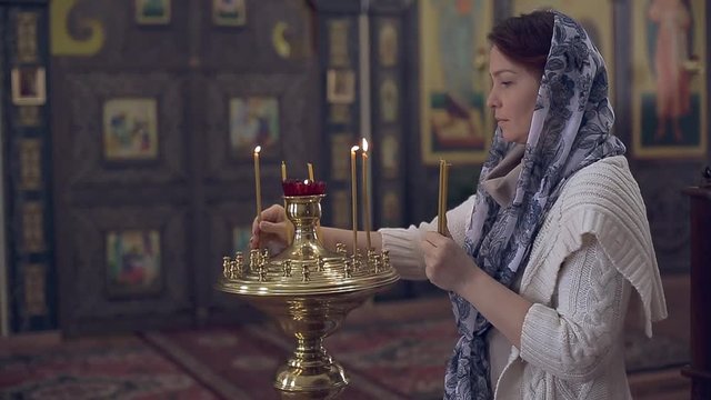 woman in the Russian Orthodox Church with red hair and a scarf on her head lights a candle and prays in front of the icon.