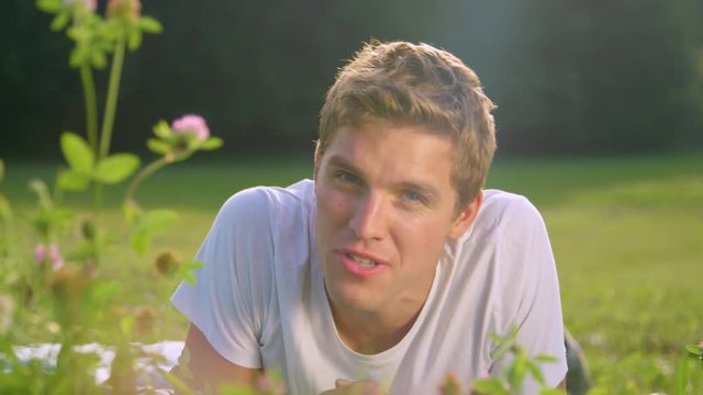 PORTRAIT, DOF: Cheerful male student filming a short video for a social media post while lying in the sunlit meadow. Smiling man waves at the camera after finishing a video call with his family.