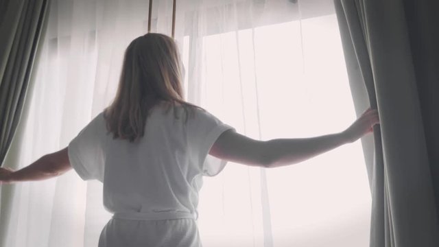 Young Caucasian Woman in White Hotel Bathrobe Unveiling Curtains and Looking Out of Window in the Morning