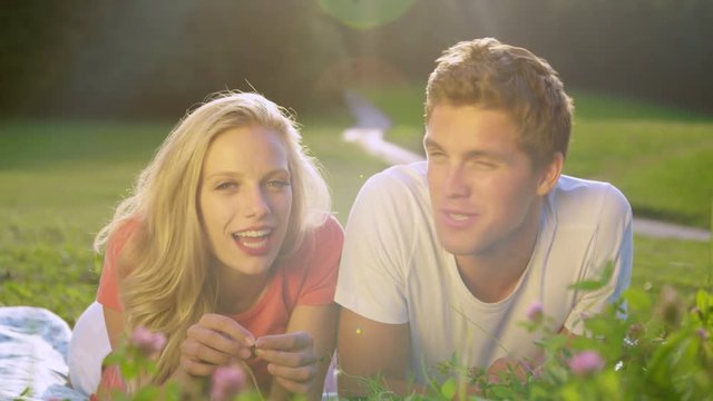 PORTRAIT SUN FLARE: Young student couple calling back home while lying in the tranquil meadow. Golden summer sunbeams shine on digital nomads filming a live video for their social media account.