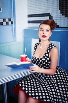 Retro (vintage) portrait of beautiful young woman sitting in cafe with book and beverage. Pin up style portrait of young woman in dress