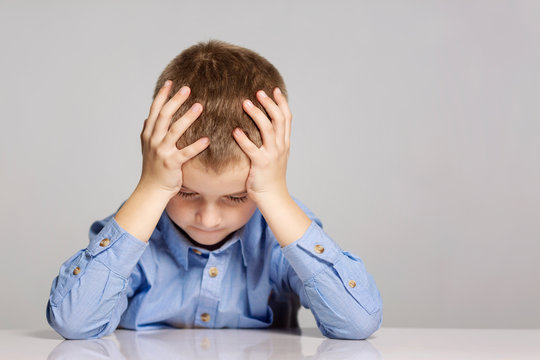 Sad Schoolboy Sitting At The Table, Holding His Head With His Hands, Gray Background