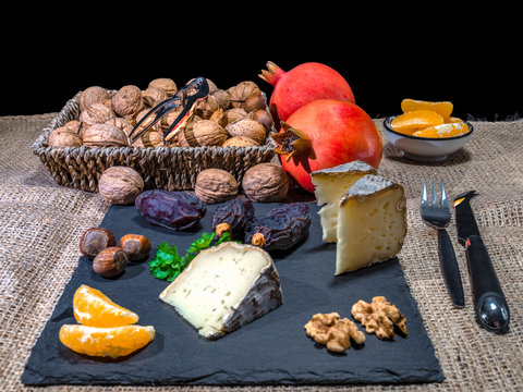 The peaces of the French cheese named Tommete des Alpes, nuts and walnuts with mandarin, date fruit and pomegranates on the natural stone plate for vegetarians as health food.