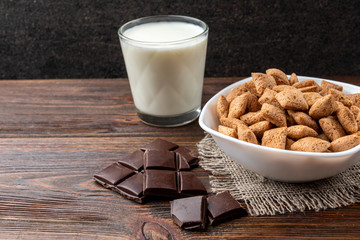 Dried breakfast pads in white bowl, milk and chocolate bar on dark wooden background.