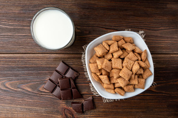Dried breakfast pads in white bowl, milk and chocolate bar on dark wooden background.