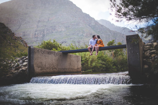 Wide angle view of a loving couple sitting on a walkway over a river stream in the mountains.