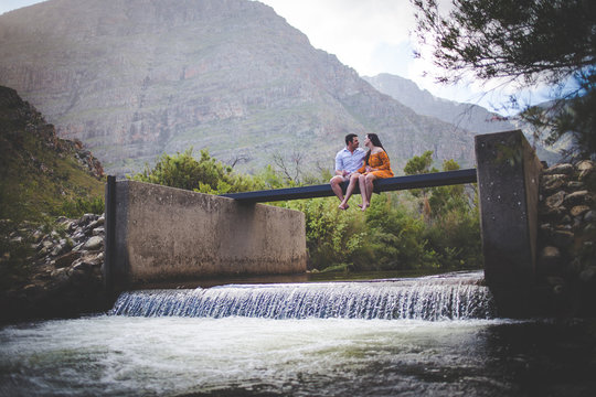 Wide angle view of a loving couple sitting on a walkway over a river stream in the mountains.