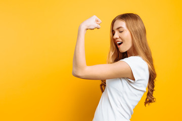 of happy girl in white t-shirt showing arm muscles on yellow background.
