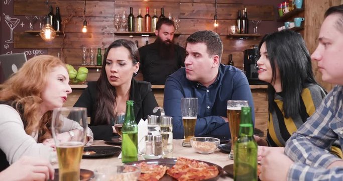 Big group of friends toasting and having fun in vintage stylish pub or restaurant. They are rising their glasses up and have fun. Hipster bearded bartender is working at the counter in the background