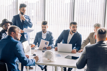 Respectable Indian employee dressed in formal suit and hat planning company costs, discussing financial strategy or marketing research during meeting at desk in office.