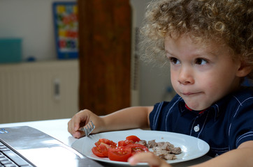 Junge sitzt mit Teller beim Essen vor einem Notebook