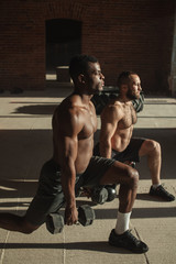 Two multiracial male crossfit sportsmen with naked torso, exercising actively in sunny morning doing lunges with weights in both hands