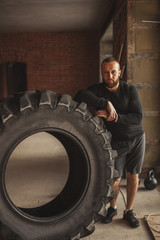 Fototapeta na wymiar Professional CrossFit trainer explains principles of training with tire during functional workouts, leaning on the huge tire in street gym