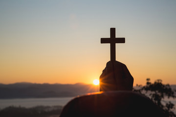 Woman hand holding holy lift of christian cross with light sunset background.