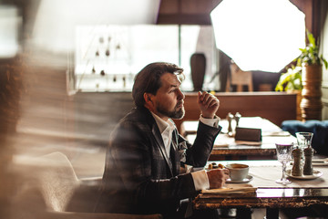 Attractive successful young businessman drinking coffee sitting at restaurant with mug and papers on table, looking through window with pensive expression