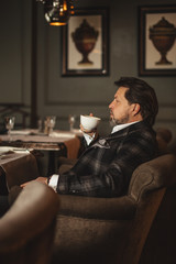 Attractive successful young businessman drinking coffee sitting at restaurant with mug and papers on table, looking through window with pensive expression