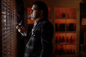 Mature businessman in stylish suit enjoying a moment of contemplation indoor in dark interior. Handsome bearded man smoking a pipe with tobacco being far away with his thoughts.