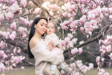 Fototapeta premium Cute Baby 6 month old Girl in Pink Outfit with Big Blue Eyes with Young Beautiful Mother at Spring, Pink Blooming Tree at the Background
