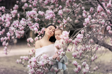 Obraz na płótnie Canvas Cute Baby 6 month old Girl in Pink Outfit with Big Blue Eyes with Young Beautiful Mother at Spring, Pink Blooming Tree at the Background